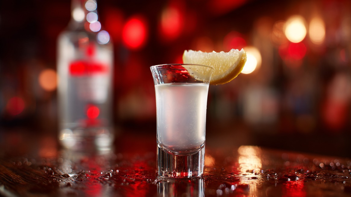 A clear vodka shot served in a small glass with a lemon wedge on the rim, set on a glossy bar counter with blurred red lights in the background