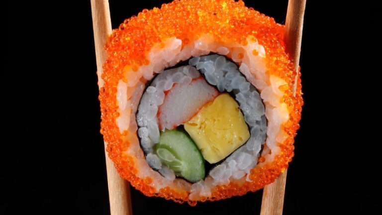 Sushi roll coated with bright orange masago roe, held between chopsticks