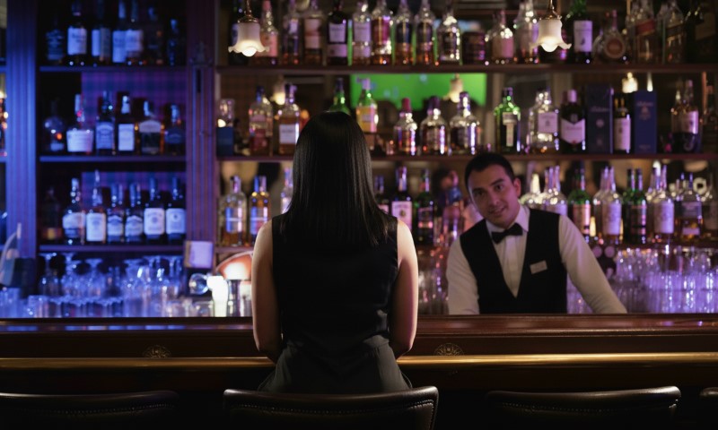 A woman with long dark hair sits alone at a dimly lit bar facing a bartender